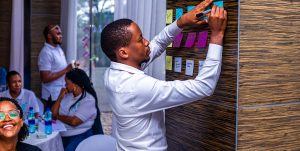 A young entrepreneur in professional clothing writes notes on a wall with marker while another group of entrepreneurs seated at a table in the background. The seated group is enjoying themselves in conversation and laughing.